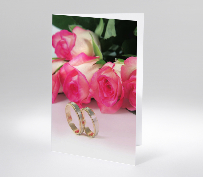 Fotokarte zur Hochzeit mit einem liegenden Blumenstrauß aus fünf pinken Rosen. Darunter zwei Eheringe.