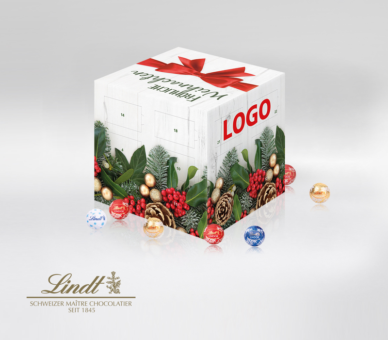 Würfelförmiger Adventskalender von Lindt mit einer fotoartigen Bordüre aus Zapfen, roten Beeren sowie Mistel- und Tannenzweigen. Auf der oberen Fläche ist eine rote Geschenkschleife und der Schriftzug Fröhliche Weihnachten.