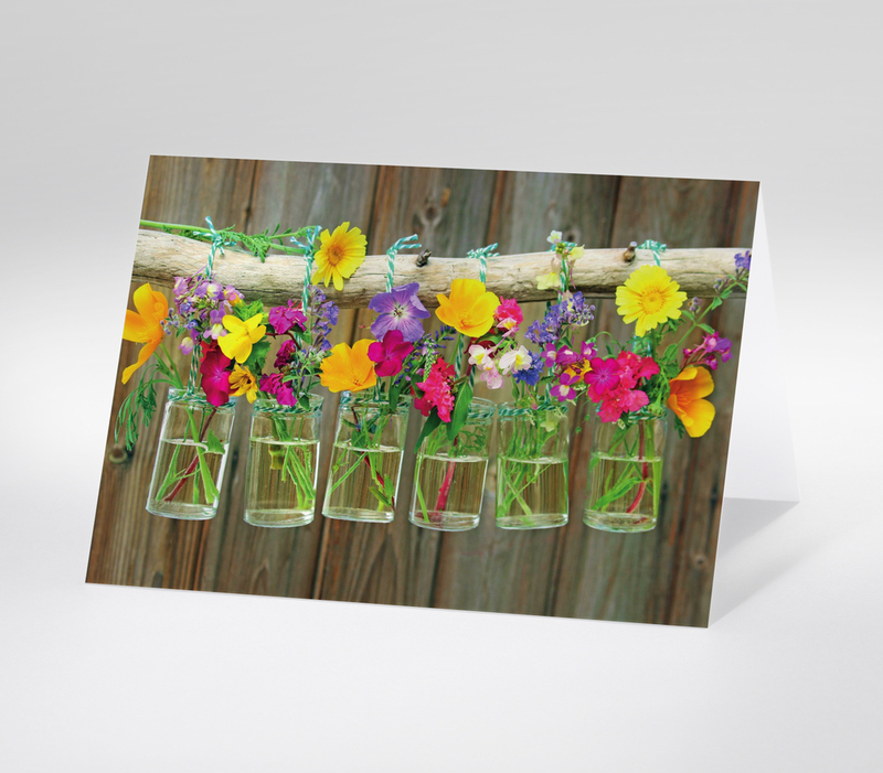 Sechs kleine Vasen mit bunten Wildblumen an Ast aufgehängt vor Holzwand