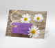 Fotokarte die drei kleine Gäseblümchenblüten auf Holz zeigt. Dabei liegt ein lila Grußzettel mit dem Text Alles Gute.